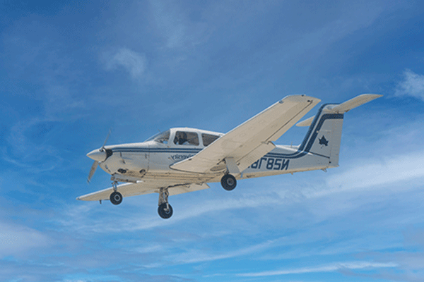 A white airplane with blue striping and blue lettering that says Indiana State University is pictured in flight against a blue sky with white clouds. N2878 can be made out on the side behind the wing. The three-wheel landing gear is down and the propeller appears to be in motion. A figure is faintly visible in the co-pilot’s seat. 