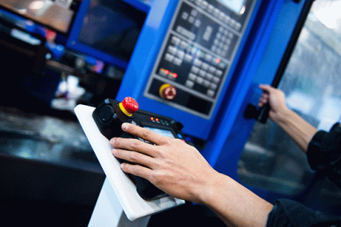 A pair of hands are shown engaging with a blue control panel. The right hand rests on a black control pad with two illuminated orange lights and a red button on a yellow base, and a few other buttons and lights on the far side. The pad sits on a white pedestal. In the background, a tall blue control panel with various buttons, lights, and a red knob is also visible. The left hand in the image rests further away on a black vertical handle attached to the control panel.